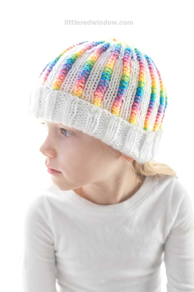 A young child wearing a charming rainbow rib hat and a white shirt gazes to the left. The soft white background enhances the bright, focused look on the child's face, highlighting the vibrant colors of their hat.