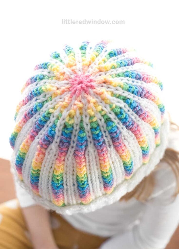 Viewed from above, a person dons a rainbow rib hat with vibrant colors and a white brim. The neutral background enhances the striking design of the knitted pattern.