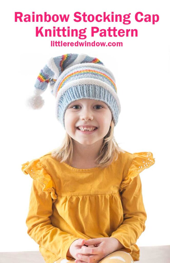 smiling girl in yellow shirt wearing a knit stocking cap with stripes of blue white and rainbow