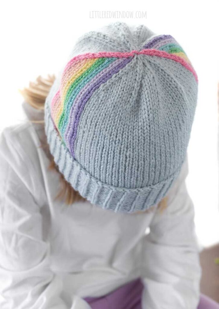 child in white shirt leaning forward towards the camera so you we can see the top of a light blue knit hat with vertical rainbow stripes twirling up towards the top in front of a white background