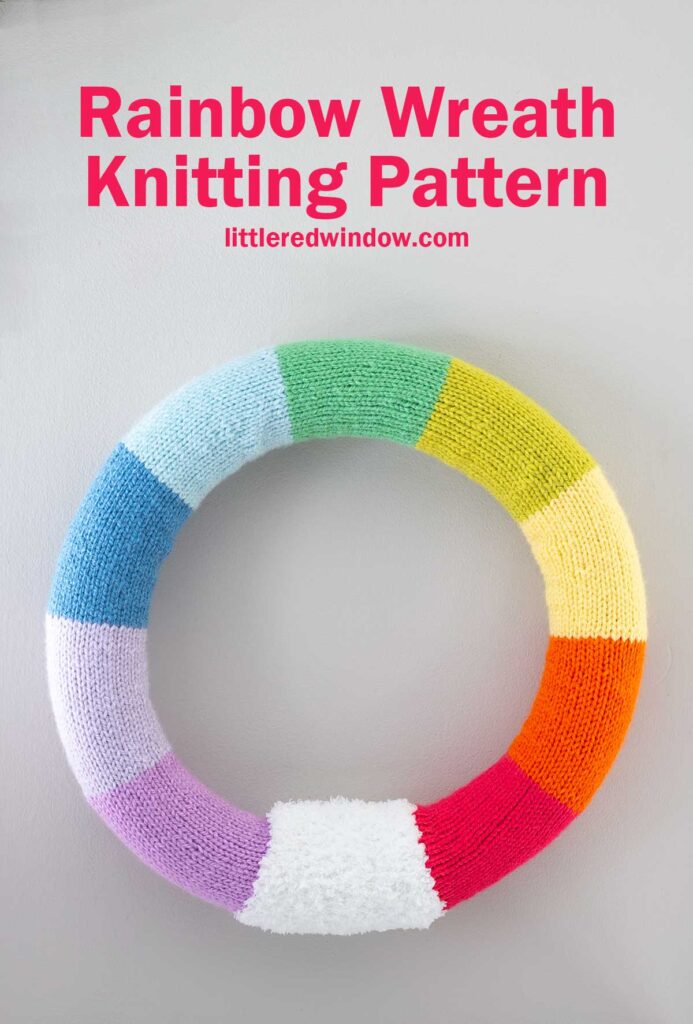 A colorful knitted wreath with rainbow segments: red, orange, yellow, green, blue, and purple. Text at the top reads 