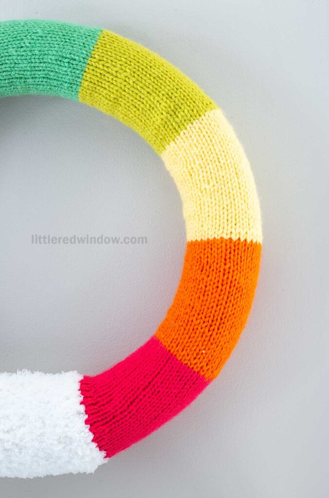 A colorful knitted wreath with sections of green, yellow, cream, orange, red, and white, against a light grey background. Visible watermark reads