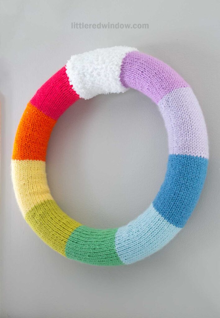 A knitted wreath featuring colorful segments in red, orange, yellow, green, blue, and purple, with a white fluffy section at the top, hanging on a light gray wall.