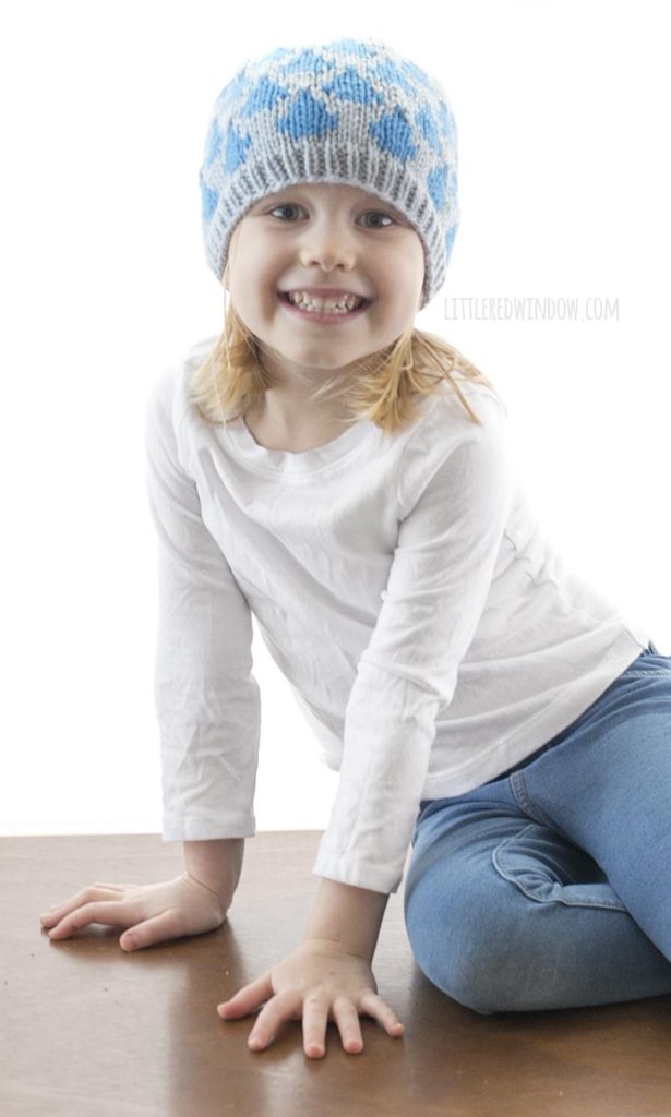 little girl in white shirt smiling and wearing blue knit hat with raindrops on it