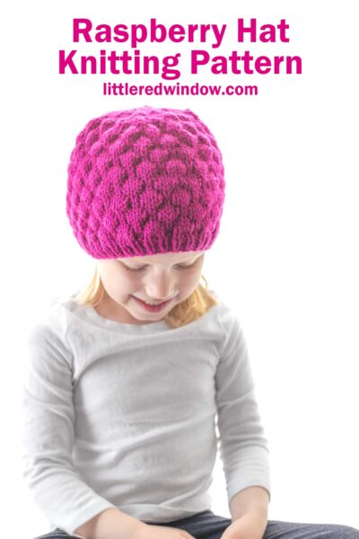 A young child wearing a bright raspberry hat smiles while looking down. The text above reads "Raspberry Hat Knitting Pattern" with the website littleredwindow.com below it. Dressed in a light gray long-sleeve shirt, the child's joy is infectious.