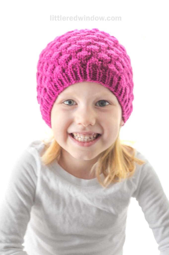 A young child with blond hair smiles brightly, wearing a vibrant, textured raspberry hat and a white long-sleeve shirt. The background is plain white.