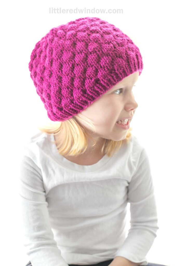 A young child with blonde hair is wearing a vibrant, textured raspberry hat and a plain white long-sleeve shirt. The child is smiling and looking to the side while standing against a bright, white background.