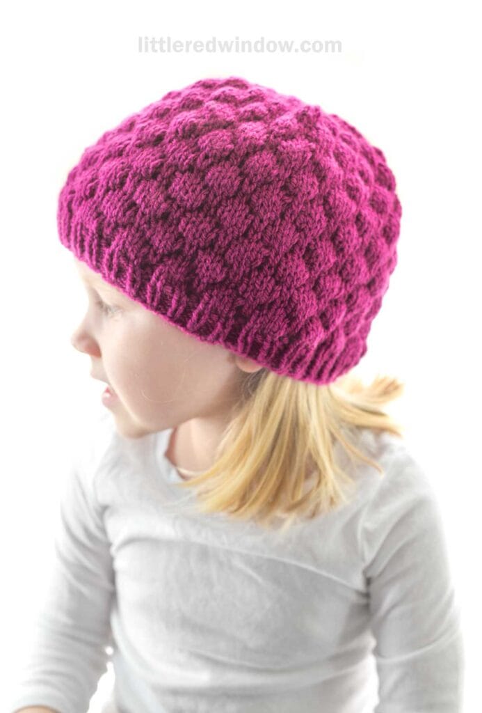 A young child with blonde hair is wearing a bright raspberry hat, textured and knit. They are dressed in a white shirt and looking to the left. The background is plain white.