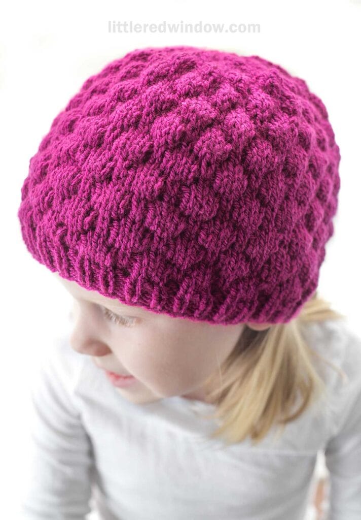 A young child wearing a textured, bright pink raspberry hat. The child is looking down and slightly to the side, and the background is softly blurred. The hat is handmade with a detailed pattern. The child is dressed in a light-colored outfit.