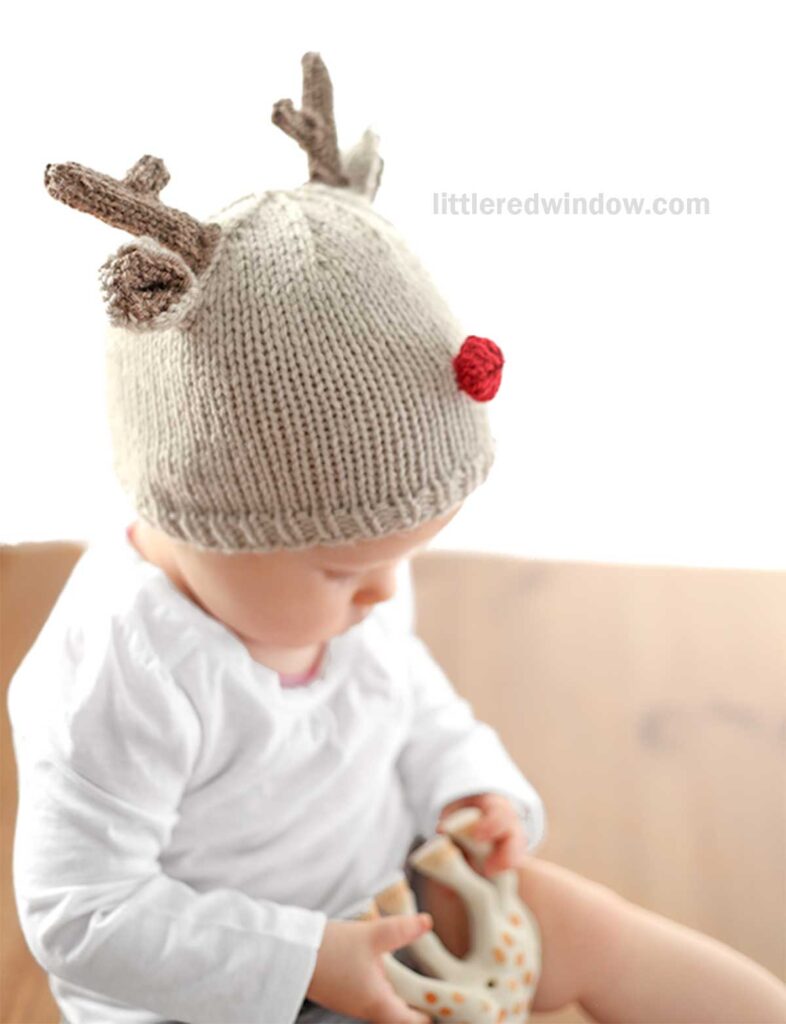 A baby sits on a wooden surface, sporting an adorable reindeer hat complete with antlers and a red nose, holding a toy. Dressed in a white shirt, the baby is set against a bright, blurred background.