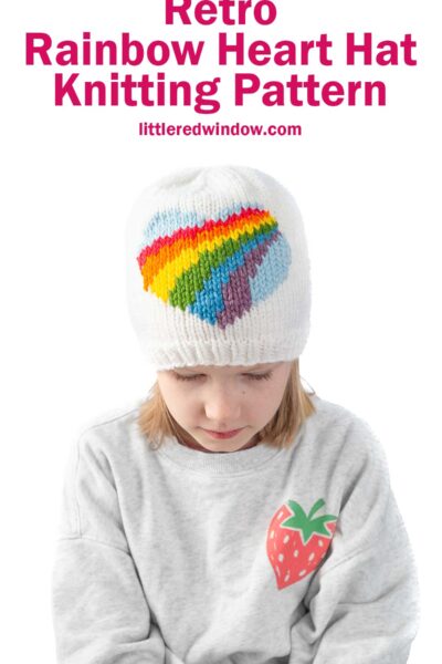 Child wearing a white knit hat with a rainbow heart design. The child is also wearing a gray sweatshirt with a strawberry patch. Text above reads "Retro Rainbow Heart Hat Knitting Pattern" and "littleredwindow.com".