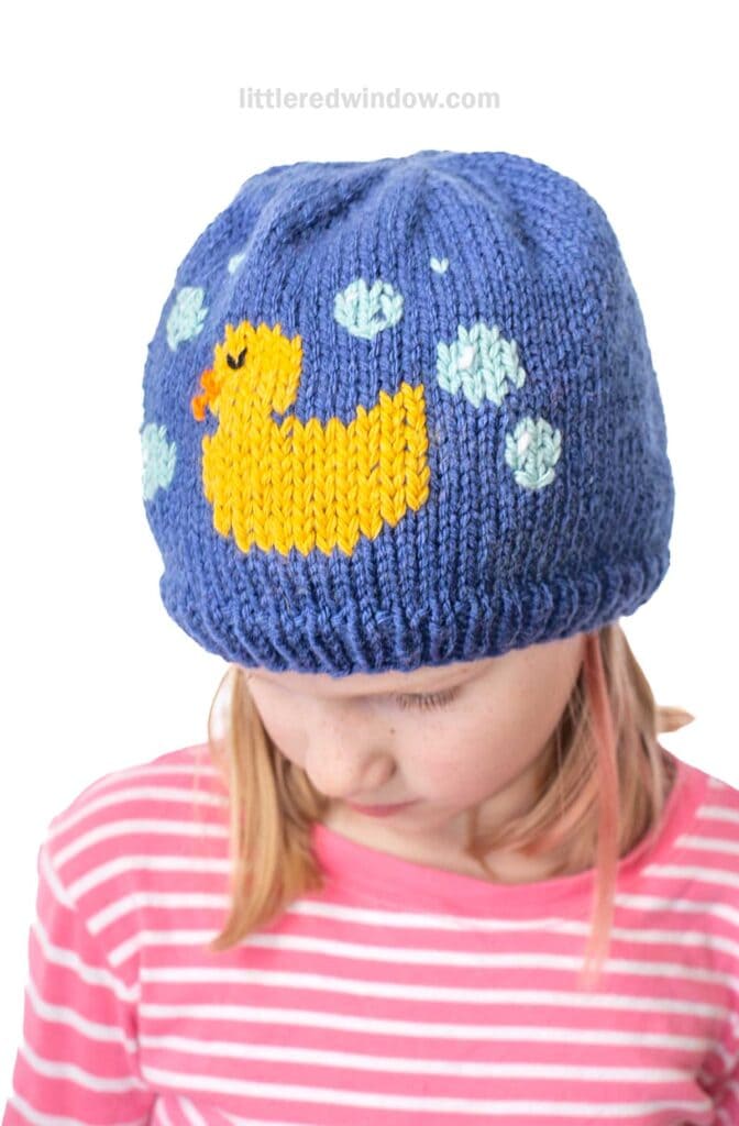 A child wearing a blue knit hat featuring a yellow duck and white bubbles pattern. They are looking down and have a pink and white striped shirt.
