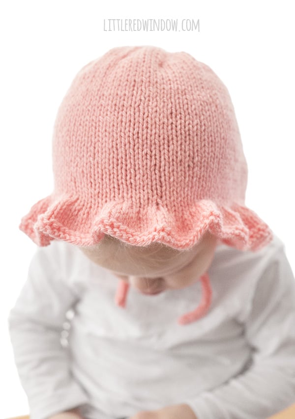 front view of chubby toddler in white shirt in front of a white background wearing a light pink ruffle bonnet knitting pattern with chin ties and looking down at their lap