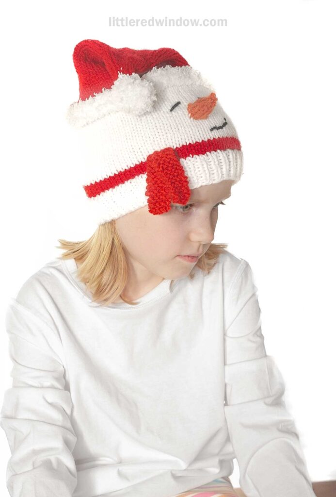 A child wearing a white sweater and a red and white knit hat designed to resemble a snowman's face. The hat features a carrot-shaped nose and button-like eyes, with a red santa hat on top. The child is looking downwards.