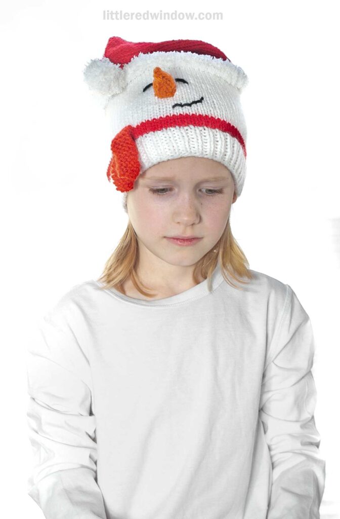 A child adorned in a charming Santa snowman hat, featuring a carrot nose and black eyes, gazes down while wearing a crisp white shirt against a simple backdrop.