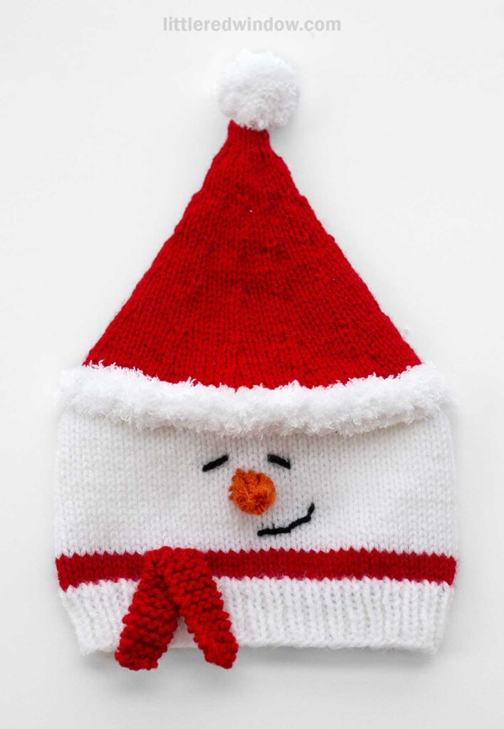 This Santa Snowman hat is a charming knitted accessory, showcasing a snowman's face complete with a carrot-like nose and a joyful smile. Topped in festive red with a fluffy white pom-pom, it's perfect for holiday cheer.
