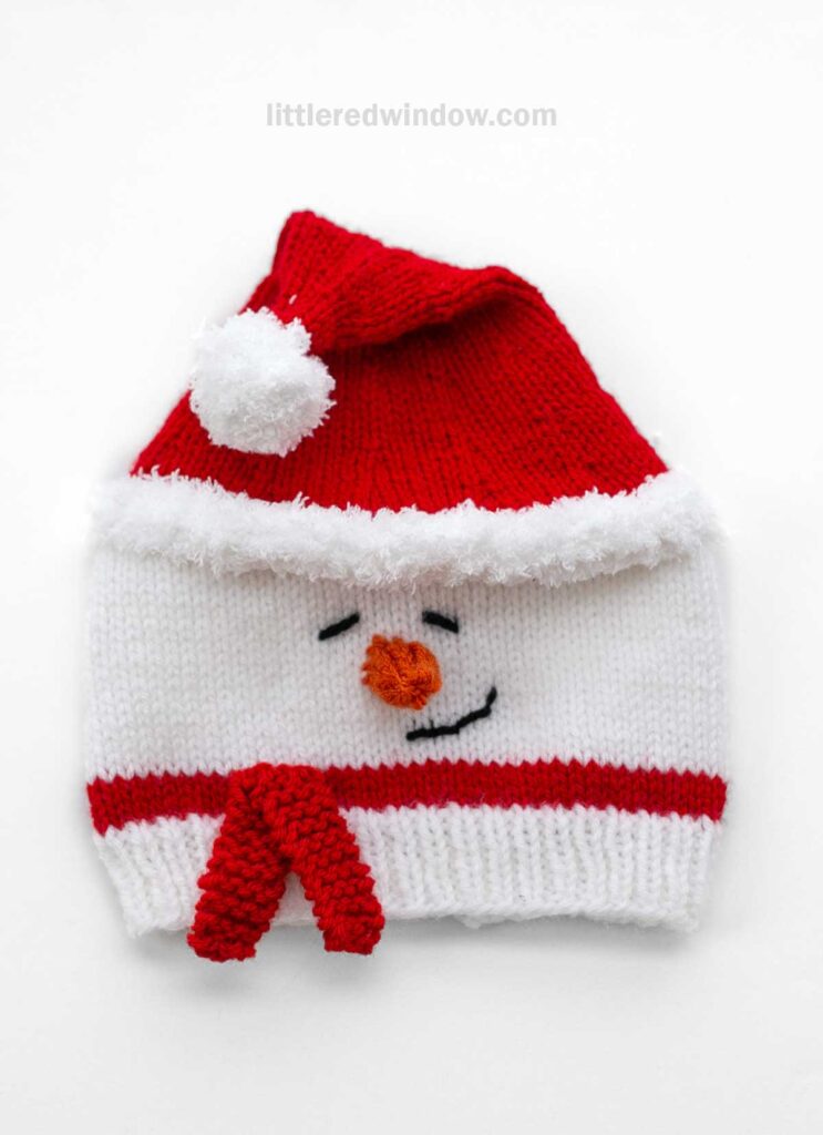 A knitted Santa snowman hat designed to delight, featuring a red top with a fluffy white pom-pom, an endearing face with an orange carrot nose, and a red scarf along the bottom.
