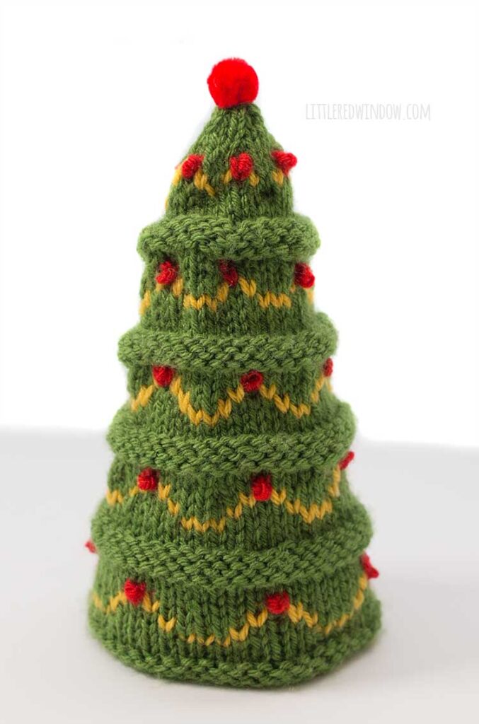 a green cone shaped knit tree with gold colored swag garlands and red ornaments and a red pom pom on top