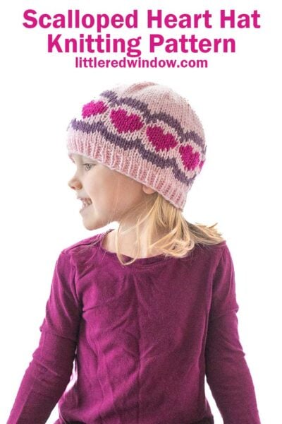The child, donning a scalloped heart hat in hues of pink and purple, gazes thoughtfully to the side. Her ensemble is completed with a long-sleeve purple shirt. The text above reads, "Scalloped Heart Hat Knitting Pattern" and "littleredwindow.com".