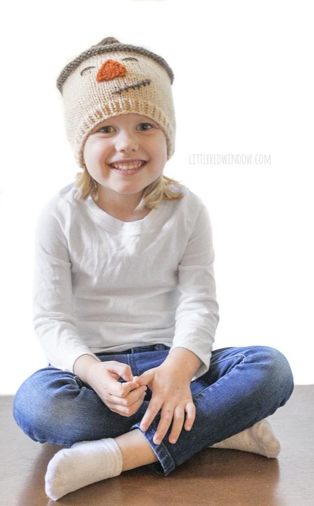 little girl in white shirt smiling and wearing knit scarecrow hat