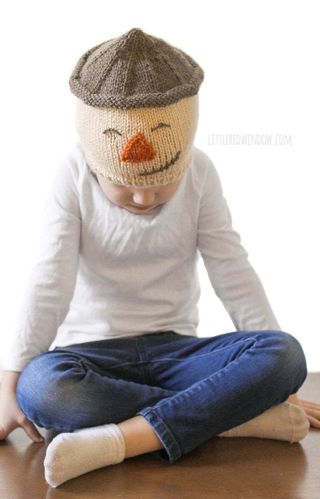 little girl looking down and wearing a tan knit hat with a scarecrow face on it