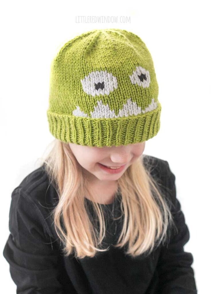 Blond girl in black shirt wearing a lime green knit hat with a monster face on the front and looking down and to the right and smiling