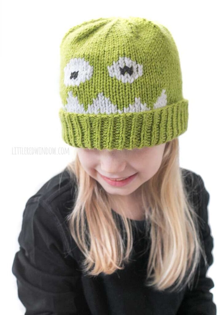 Blond girl in black shirt wearing a lime green knit hat with a monster face on the front and looking down and to the left and smiling