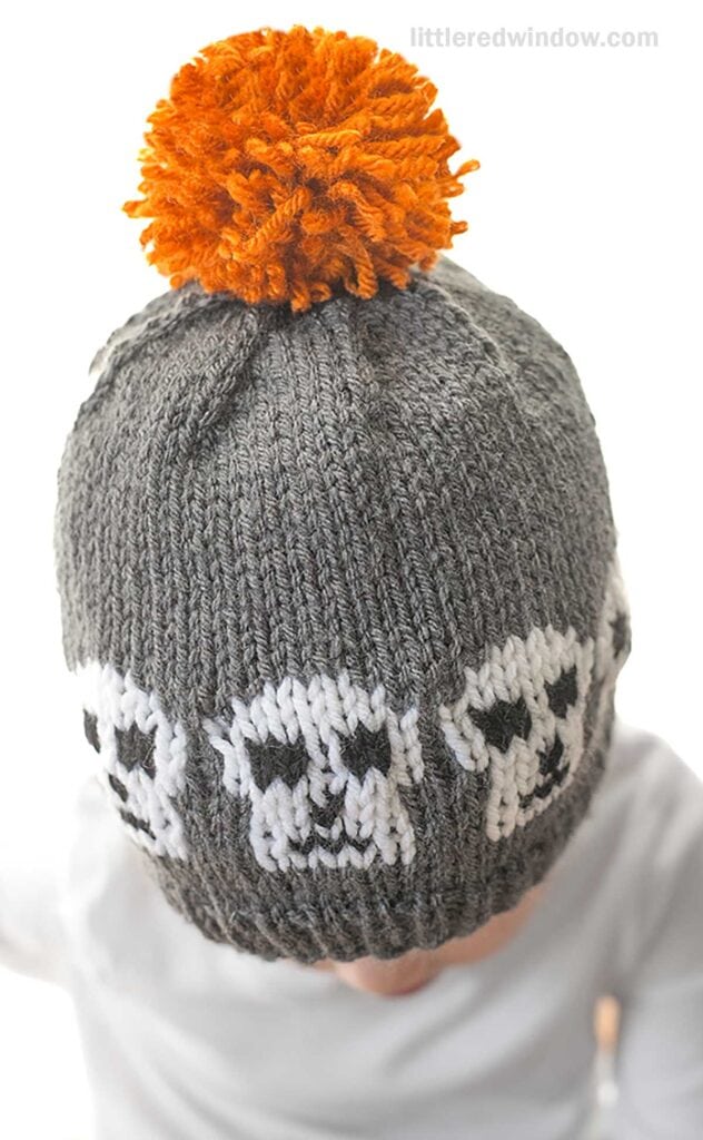 A child wearing a knitted gray beanie with white skull patterns and an orange pom-pom on top. The child’s face is not visible, and the background is white.