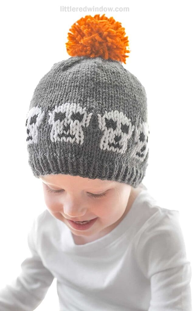A young child wearing a gray knit hat with a pattern of white skulls and a fluffy orange pom-pom on top. The child is smiling and looking down, clad in a white long-sleeved shirt.