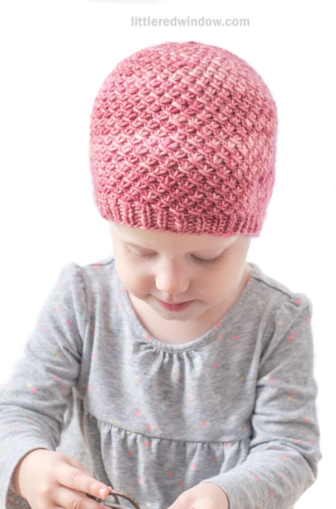 Baby in gray shirt wearing a medium pink hand dyed knit hat with a swirling slip knit twist pattern looking down at their lap in front of a white background