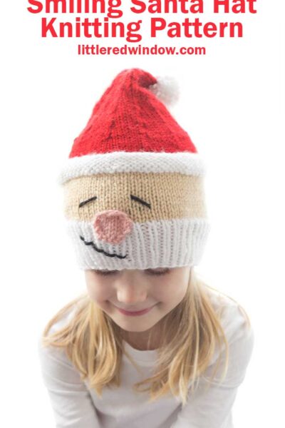Girl in white shirt wearing a knit hat that looks like Santa's head complete with white beard rosy nose and red stocking cap on top she's looking down and smiling
