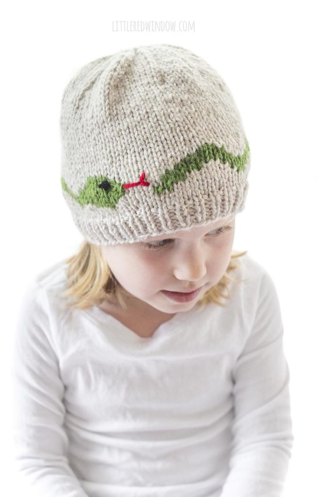girl in white shirt wearing tan knit hat with green snake with red forked tongue knit around the center she is looking down and to the right