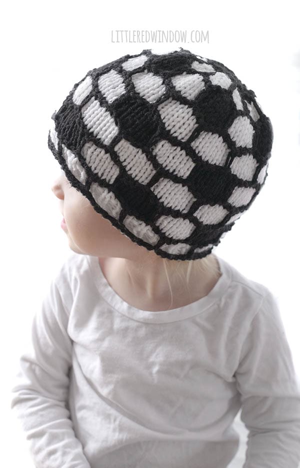 little girl in white shirt wearing black and white soccer ball hat and looking off to the left