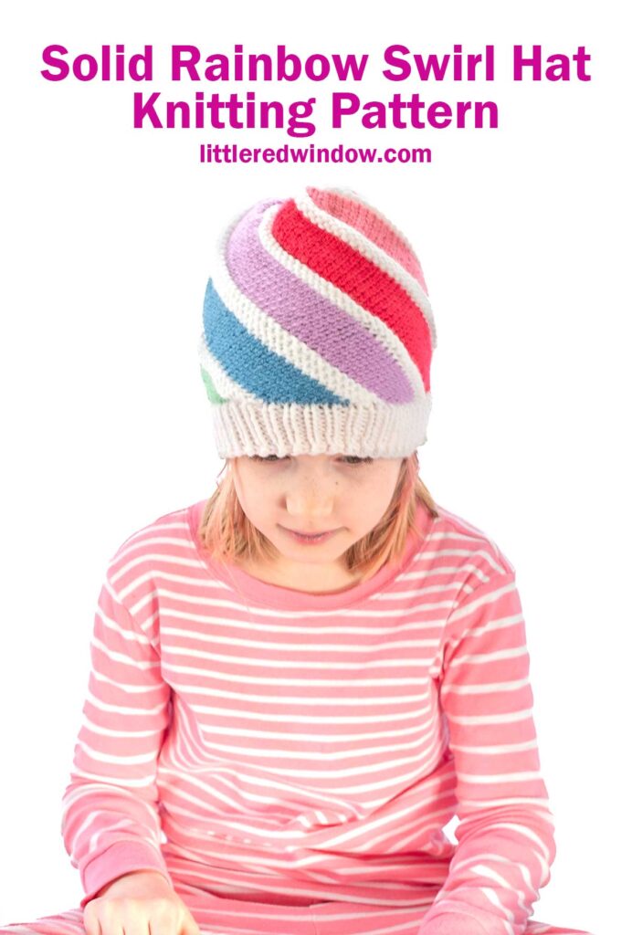 A child wearing a colorful knitted hat with swirling rainbow stripes sits against a white background. They are dressed in a pink and white striped top. The text above reads,