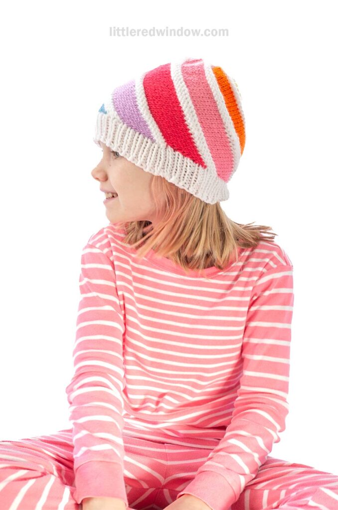 A child wearing pink and white striped pajamas is smiling while looking to the side. They have a knit hat with colorful stripes in red, pink, orange, and blue. The background is plain white.