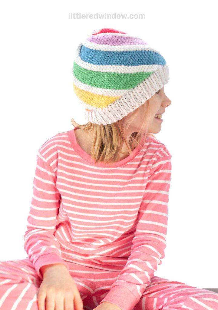 A child wearing pink and white striped pajamas and a colorful, striped knit hat looks to the side. The background is white, and the atmosphere is cheerful and relaxed.