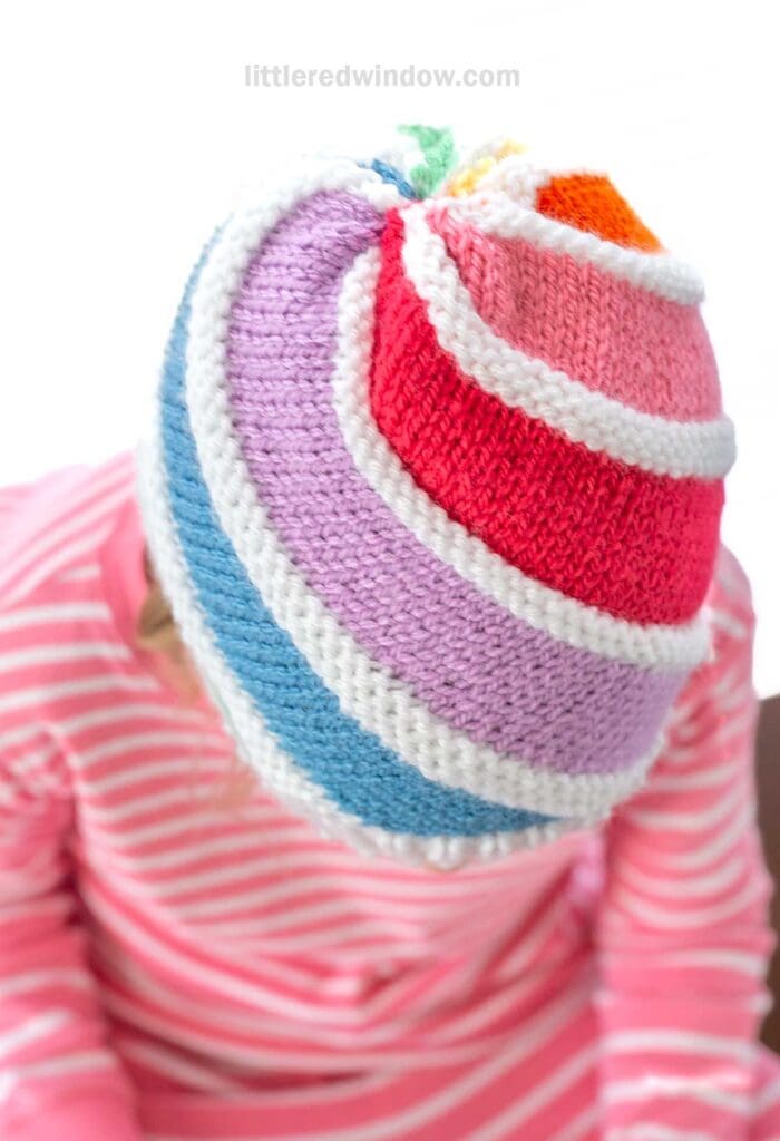 A child wearing a colorful knitted hat with rainbow stripes of pink, red, purple, blue, white, and orange. The child is also wearing a pink and white striped shirt. The background is white.