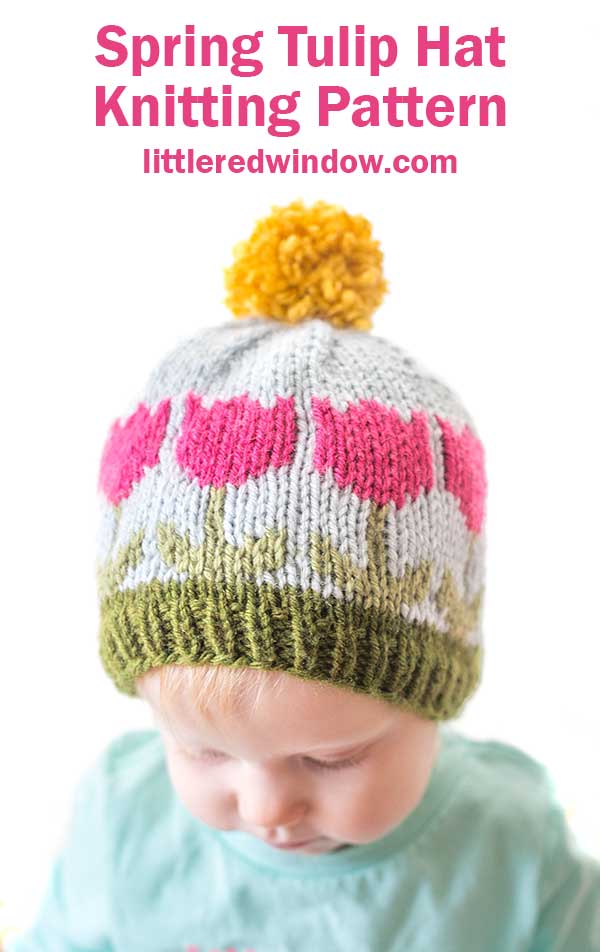 A baby wearing a knitted hat with pink tulip patterns, green leaves, and a yellow pom-pom on top. The text above reads,