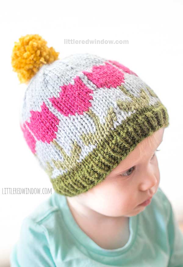 A baby wearing a knitted hat with pink tulip patterns on a light blue background. The hat features a green band at the bottom and a large, yellow pom-pom on top. The baby is dressed in a turquoise shirt.