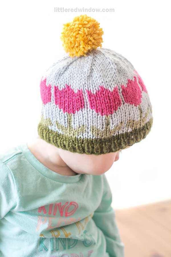 A toddler wearing a green shirt and a knitted hat with pink tulips, a yellow pom-pom on top, and green edging sits facing slightly away. The background is bright and softly blurred.