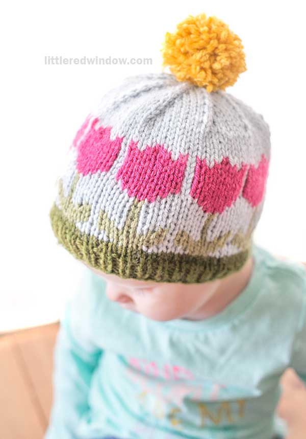 Child wearing a knitted hat with pink tulip design, a green band at the bottom, and a yellow pom-pom on top. The hat's pattern includes gray and green colors. The child is wearing a light green top.