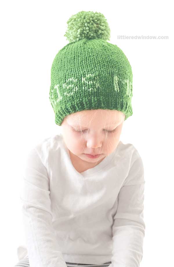 A young child is dressed in a white long-sleeve shirt and a green knit St. Patrick's Day hat adorned with a pom-pom, text knitted into the festive accessory. The child looks down thoughtfully against a plain white background.