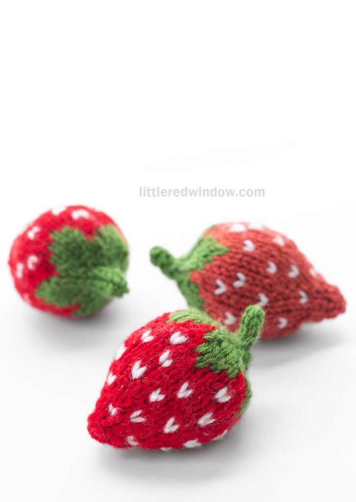 two red and one light red knit strawberries with green knit leaves and stem sitting on a white table