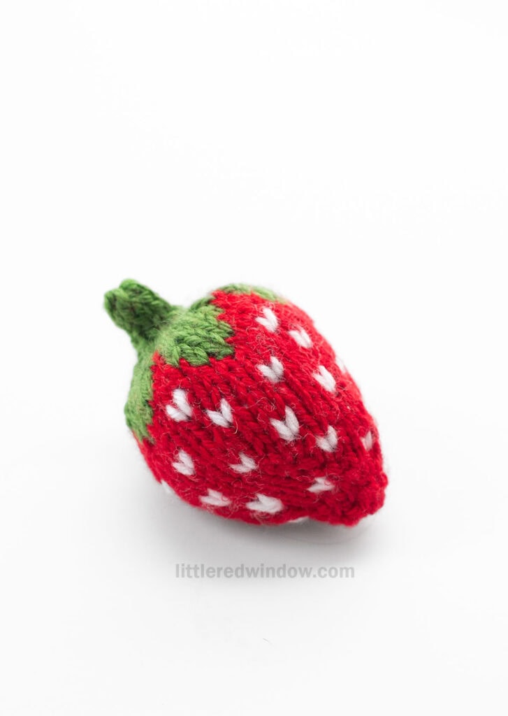 single knit red strawberry with white seed stitches and knit green leaves and stem on top sitting on a white tabletop