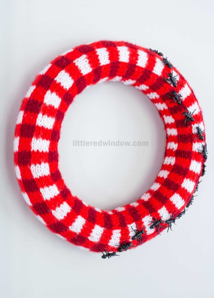 A red and white plaid knitted wreath hangs on a wall, decorated with a group of small black plastic ants crawling on the lower right side.