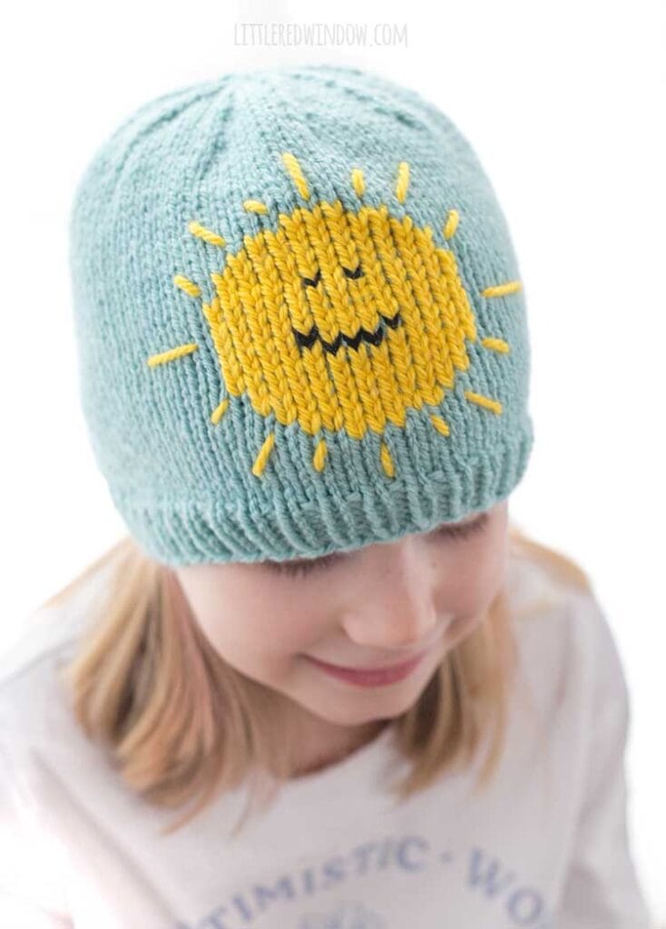 view from slightly above of smiling child in white shirt with blonde hair wearing a sky blue knit hat with a bright yellow smiling sunshine on the front looking down to the right