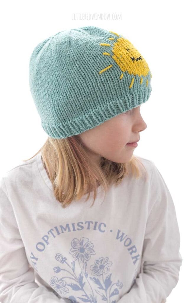 side view of child in white shirt wearing sunshine hat knitting pattern in sky blue and yellow looking off to the right