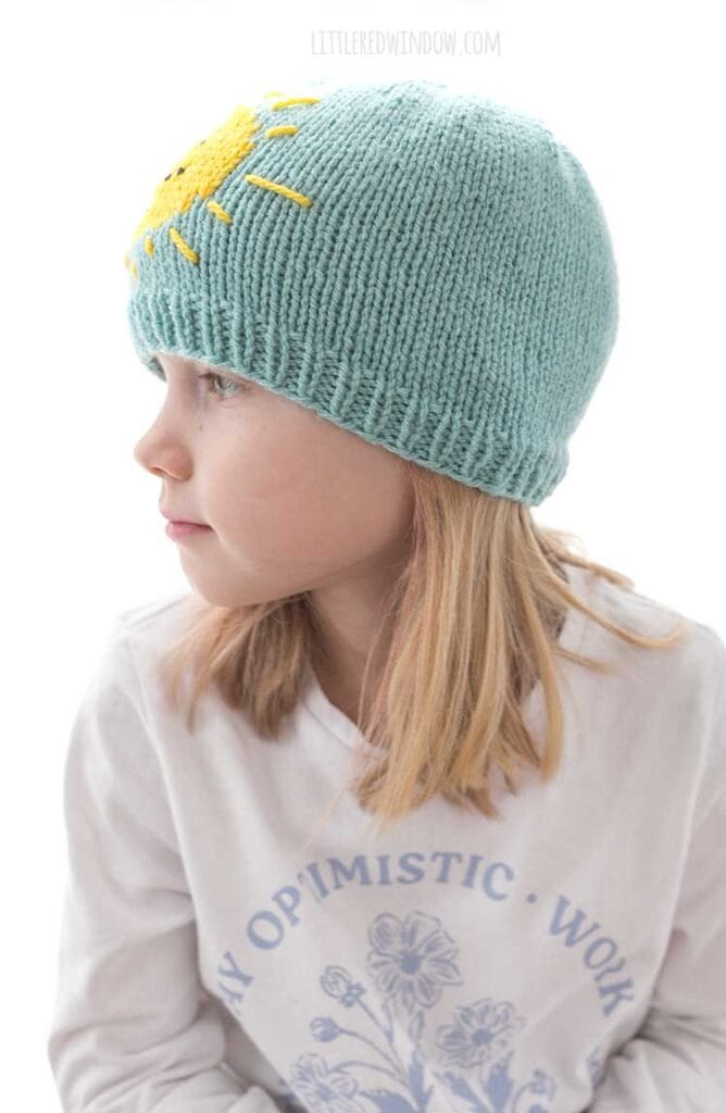 side view of child in white shirt wearing sunshine hat knitting pattern in sky blue and yellow looking off to the left