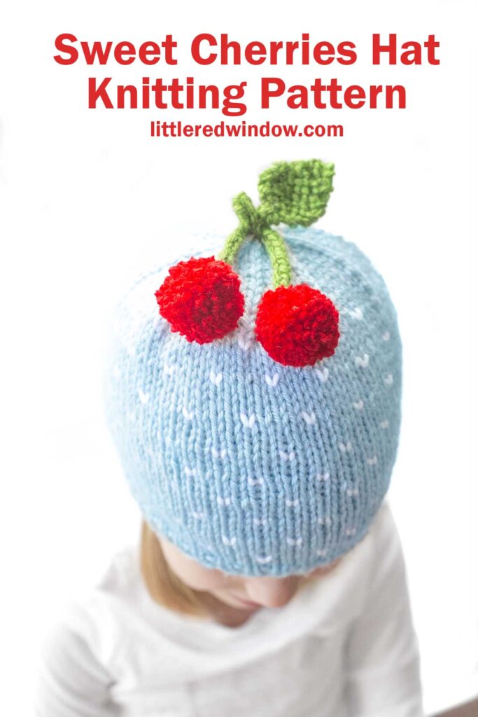 A young child wearing a light blue knitted cherry hat adorned with two red pom-pom cherries and green knitted leaves on top. Text above reads, 