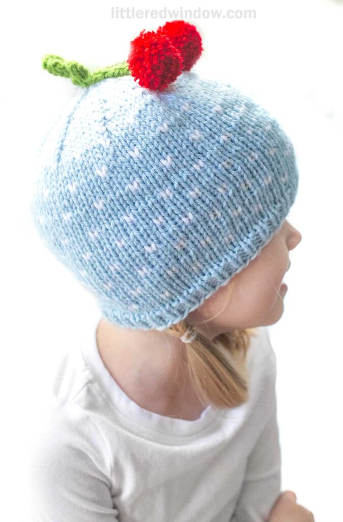 A young child wearing a light blue knit cherry hat adorned with white polka dots sits facing slightly to the side. The hat has a red pom-pom resembling a cherry with a green knit stem on top. The child wears a white shirt, and their hair is styled in low pigtails.